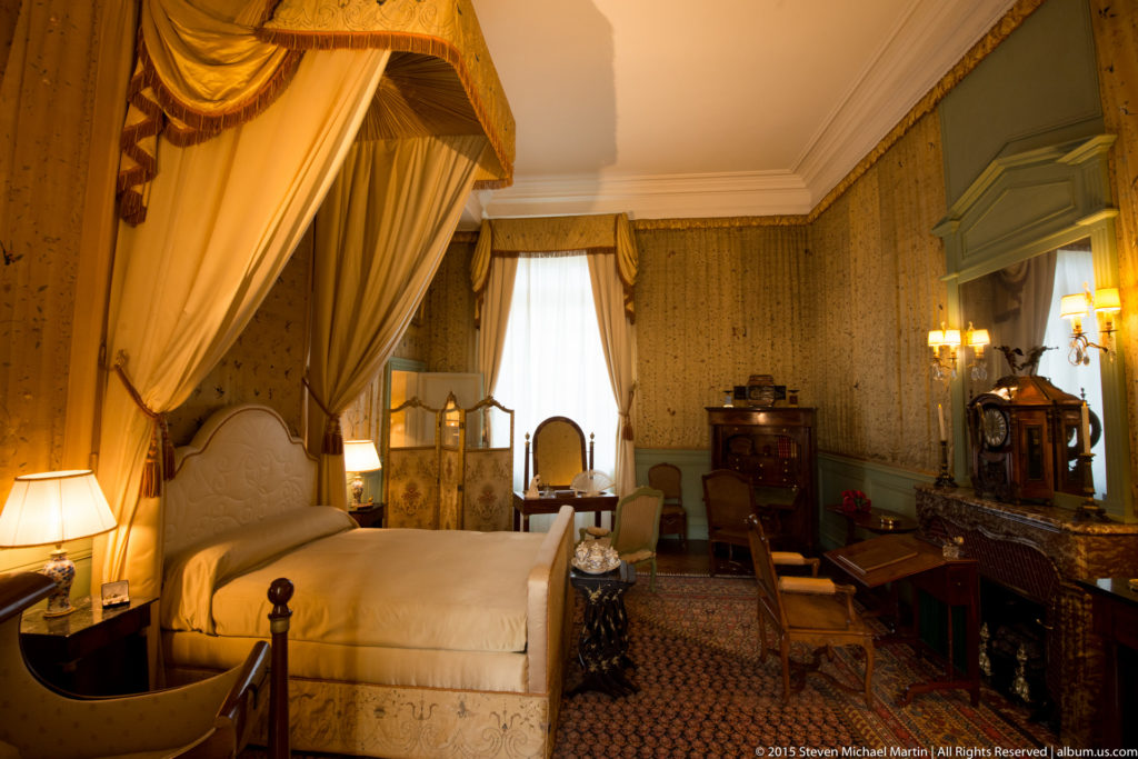 Historic château bedchamber with a pale canopied bed, patterned walls, desk, lamps, cabinetry, and heavy window drapery.