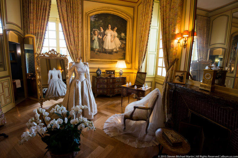 Warm salon with pale dresses on mannequins, upholstered seating, cabinets, fireplace, portrait, curtained windows, and polished wood floor.