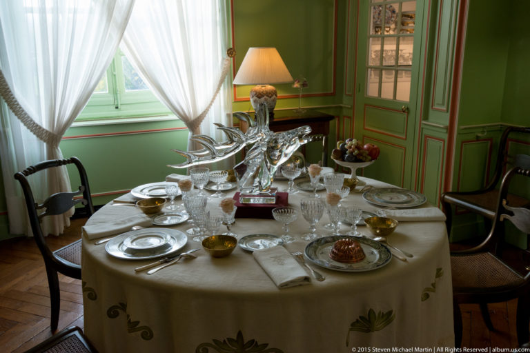 Round table set with white linen, plates, crystal glassware, and serving pieces in a green-paneled room with sheer curtains.