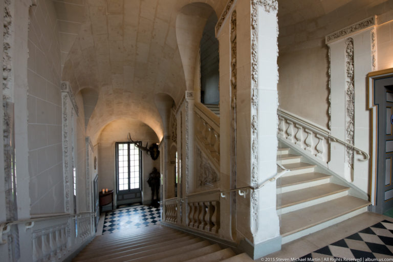 Stone staircase descends toward a checkerboard floor and tall glazed door, framed by pale vaults, carved balustrades, and relief ornament.