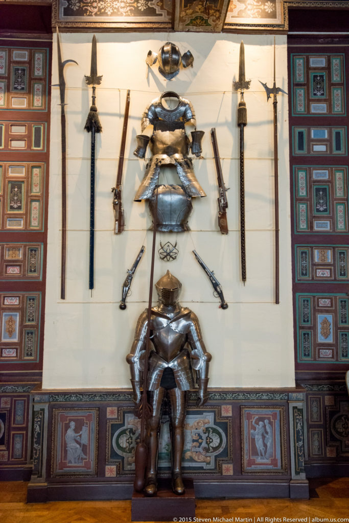 Wall display of armor, swords, polearms, and other weapons framed by richly decorated painted and carved panels.