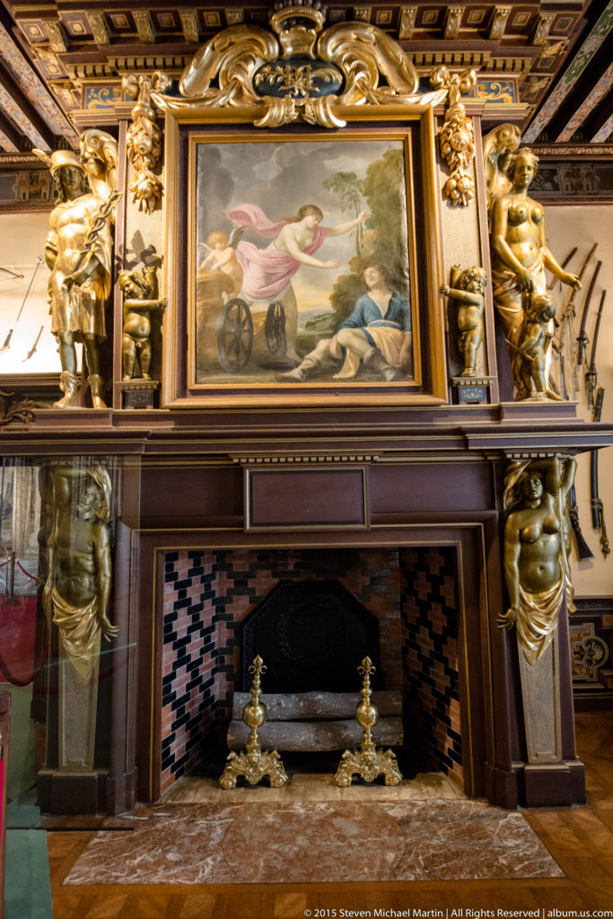 Ornate château fireplace with gilded sculptural supports, carved mantel, painting above, patterned firebox tiles, and brass hearth fittings.