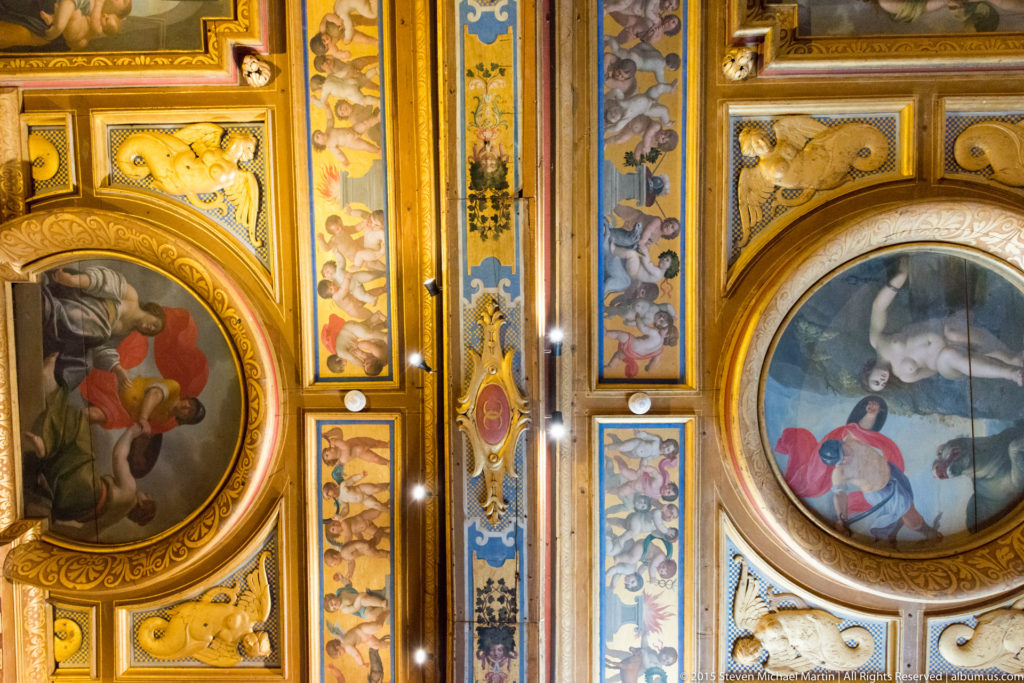 Upward view of a richly decorated ceiling with gilded moldings, colorful painted panels, and oval figural scenes.