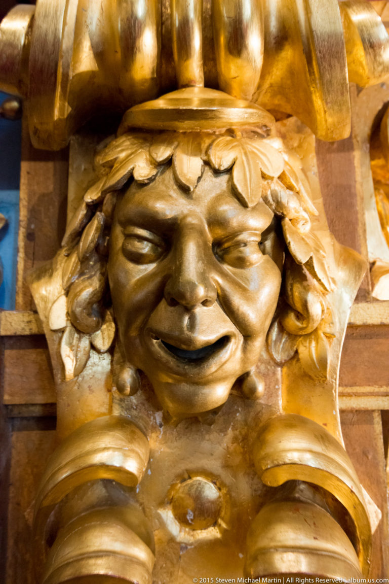 Close-up of a gilded carved mascaron with smiling face, leaf-like headdress, and surrounding ornamental scrollwork.