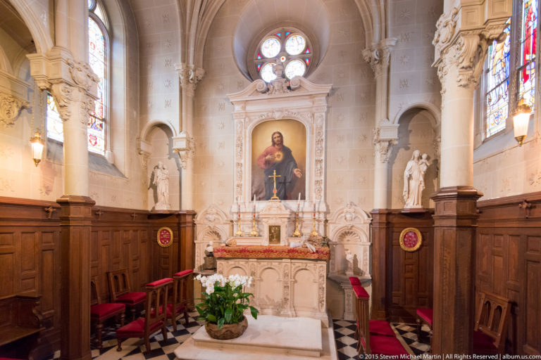 Château chapel with altar, sacred painting, circular stained glass, carved figures, tall windows, wood paneling, and red chairs.