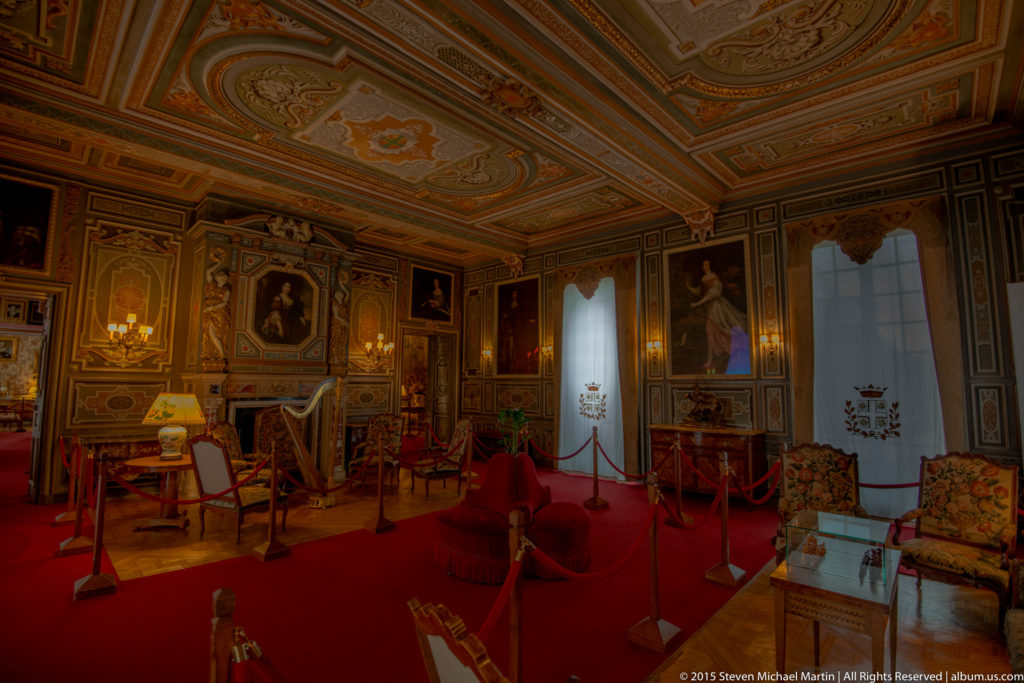 Formal château reception room with red carpet, upholstered chairs, small tables, framed portraits, patterned paneling, and visitor ropes.