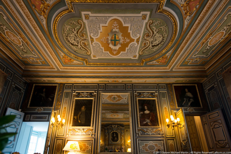 Upward interior view with ornate painted ceiling, central heraldic motif, framed portraits, dark paneling, and warm wall lights.