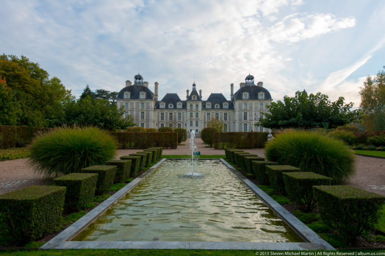 Formal garden with long rectangular water basin, clipped hedges, ornamental grasses, and Château de Cheverny centered in the distance.