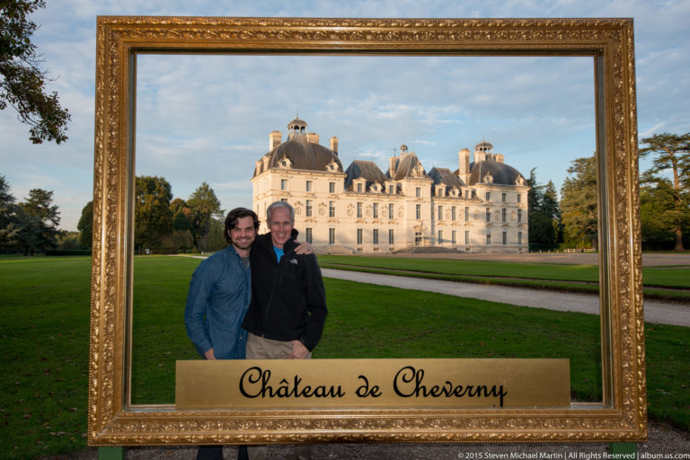 Two visitors pose inside an oversized gilded frame labeled Château de Cheverny, with the château and lawn visible behind them.
