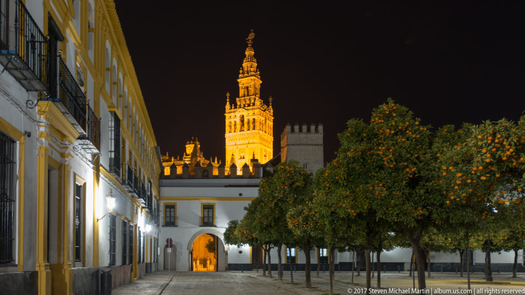 Night street lined with orange trees and historic buildings, with an illuminated archway and the Giralda glowing above in the distance.