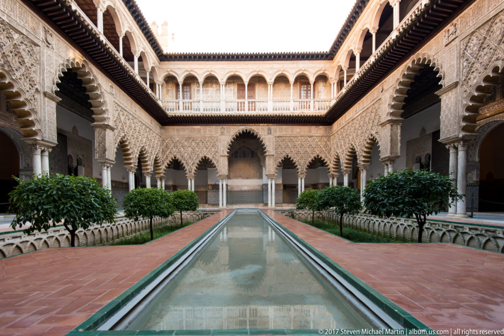 Low view along the Patio de las Doncellas reflecting pool, flanked by terracotta paving, gardens, citrus trees, and ornate arcades.