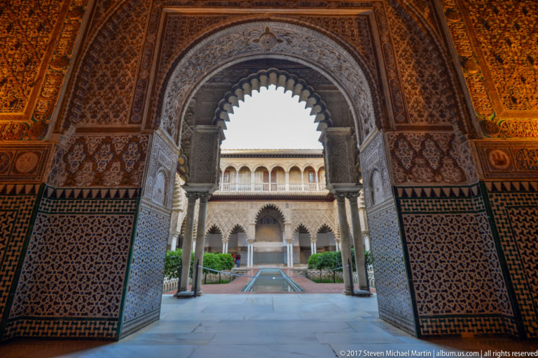 Scalloped, plaster-carved arch and tiled walls frame a view of the Patio de las Doncellas reflecting pool and arcades.