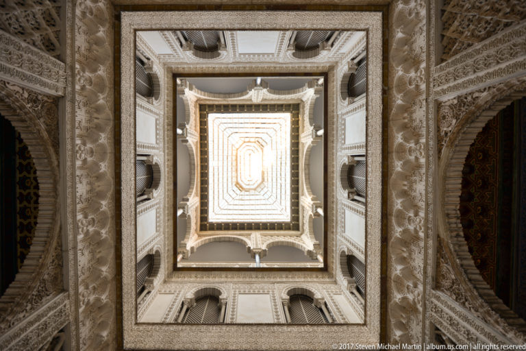 Straight-up view through stacked arcades and balconies of the Patio de las Muñecas toward a bright rectangular opening overhead.