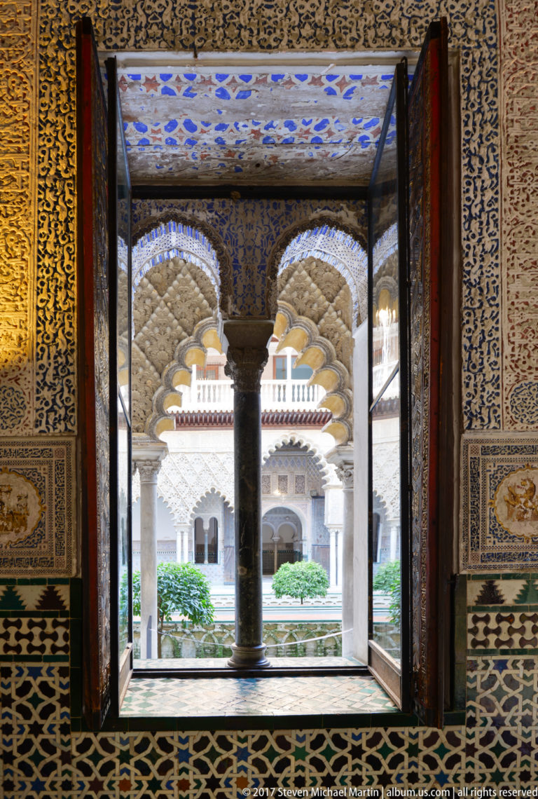 Dark central column and ornate tiled opening frame the small arcaded Patio de las Muñecas beyond.