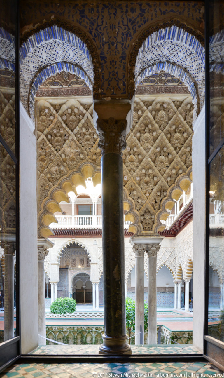 Dark column beneath patterned arches overlooks white columns, upper gallery, and greenery in the Patio de las Muñecas.