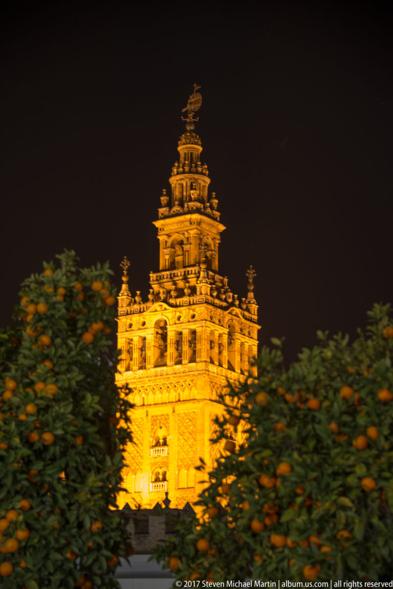 Illuminated upper Giralda tower rising above orange trees at night, with ornate arcades and belfry glowing against a black sky.