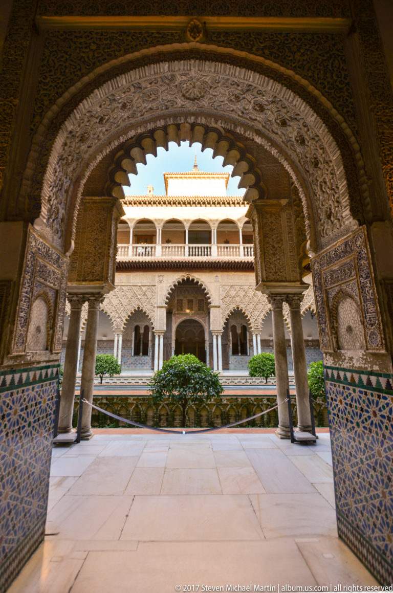 Broad carved arch with blue-and-white tile dado frames the compact Patio de las Muñecas, its central tree and opposite gallery.