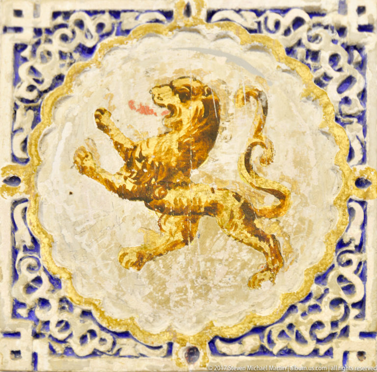 Close-up of a glazed ceramic tile with an ochre rampant lion in a pale medallion surrounded by blue interlace.