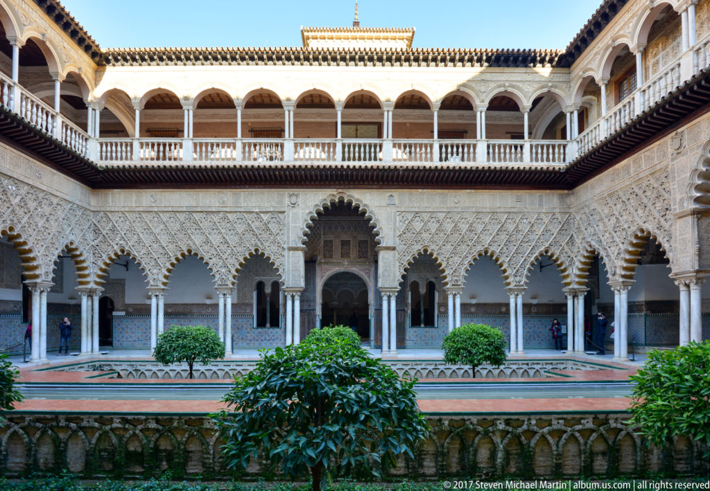 Citrus trees stand before a long arcade of slender columns, multilobed arches, carved plaster, and an upper gallery at the Alcázar.
