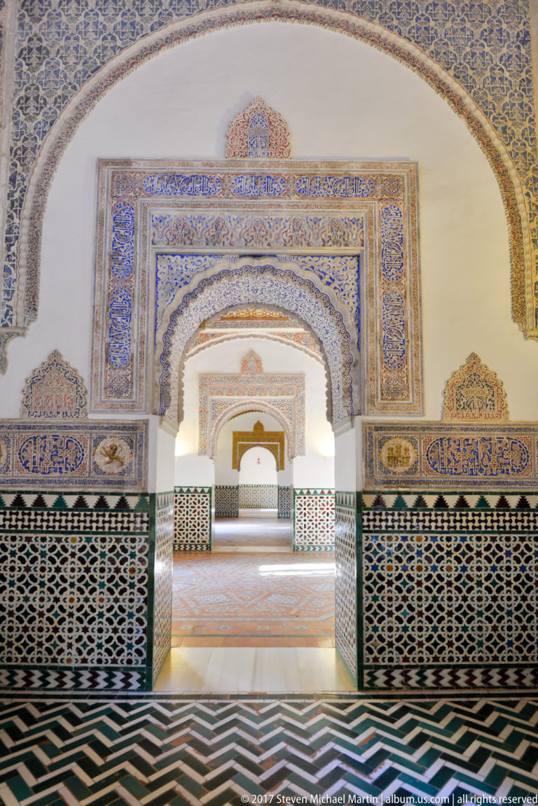 Several ornamented horseshoe arches align through tiled palace rooms, with patterned dados and terracotta floors receding toward a distant doorway.