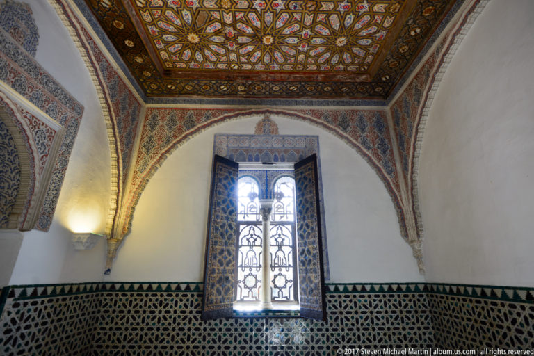 Arched window in a white palace wall above dark geometric tile, with richly painted and gilded ceiling ornament overhead.