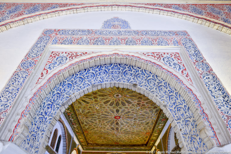 Upward view through a blue-and-red ornamented arch toward a star-patterned wooden ceiling with carved plaster surrounding the opening.