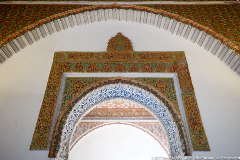 Polychrome horseshoe arch with blue inner decoration and layered red, green, and gold ornamental bands beneath a larger white arch.