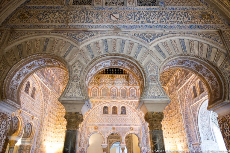 Three horseshoe arches on dark columns beneath Arabic calligraphy and dense plaster ornament in the Hall of Ambassadors.
