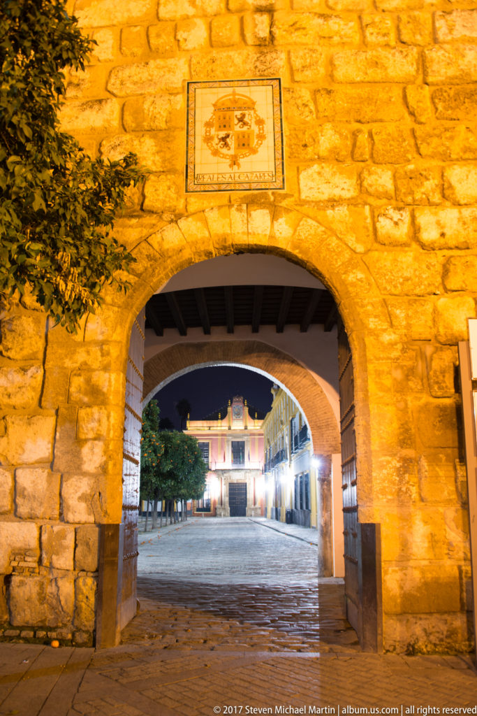 Illuminated stone-and-brick archway in the Alcázar perimeter wall opens toward a paved square at dusk.
