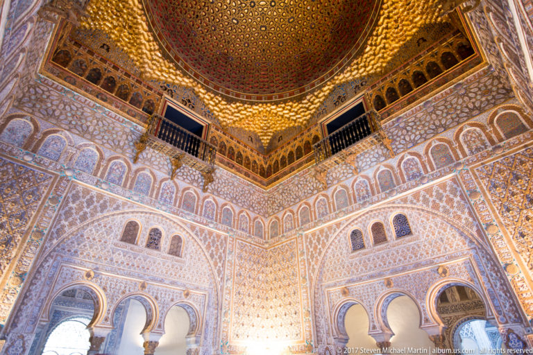 Steep corner view of the Hall of Ambassadors showing scalloped arches, carved walls, blind arcades, and the glowing gilded dome above.