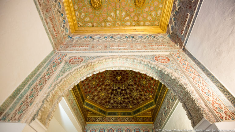 Upward view of a decorated arch between two geometric wooden ceilings, with red-and-blue painted plaster around the opening.