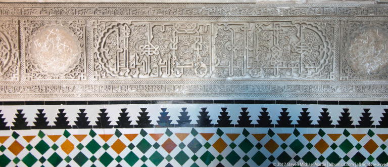 Carved pale plaster panels with calligraphic ornament above a dado of green, orange, black, white, and blue geometric tiles.