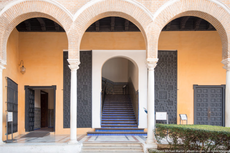 Small ochre-walled courtyard with brick arches on white columns, dark paneled doors, central staircase, marble paving, and clipped shrubs.