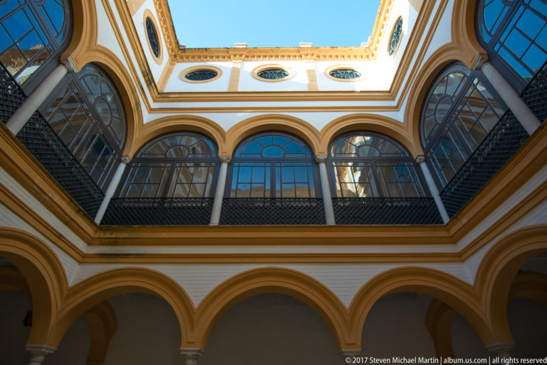 Upward view of a two-story courtyard with ochre-trimmed arches, slender columns, large glazed windows, circular openings, and blue sky above.