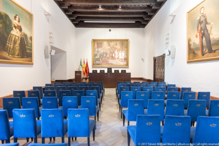 Formal white-walled meeting room with rows of blue chairs, timber ceiling, framed paintings, flags, and a front dais.
