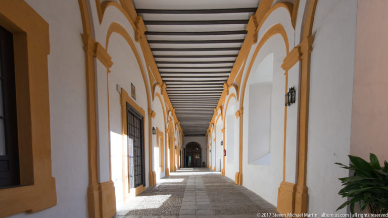 Long white corridor with ochre-trimmed arches, dark timber ceiling, stone paving, windows, wall lamps, and deep perspective.