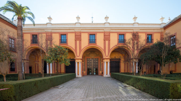 Rose-and-ochre two-story palace façade with ground-floor arches, central doorway, clipped hedges, paved path, and citrus trees.