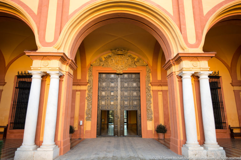Large paneled doorway beneath an ochre arch and decorative relief, framed by paired pale columns and black iron window grilles.