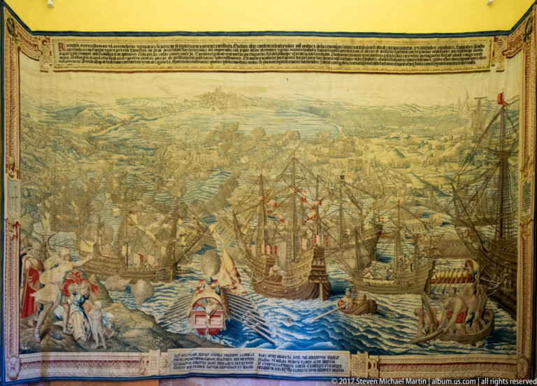 Large tapestry showing numerous sailing ships and boats in a naval battle before a distant coast, with figures, heraldry, and inscriptions.