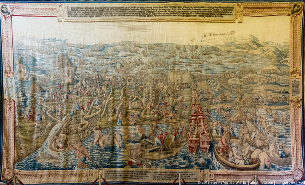 Panoramic tapestry filled with soldiers, boats, fortified settlements, waterways, and distant hills, bordered by woven inscriptions.
