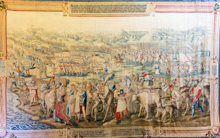 Large tapestry with mounted figures, soldiers, horses, attendants, shoreline activity, and distant troops spread across a broad landscape.