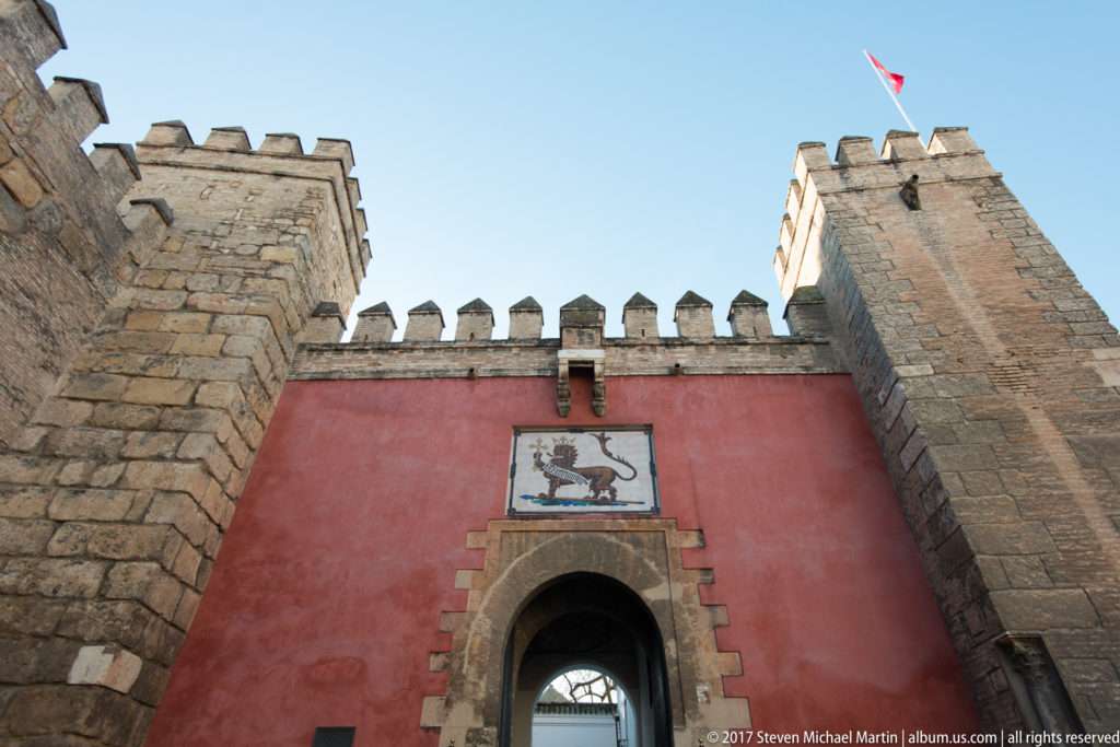 Red crenellated wall and stone towers frame the pointed Lion Gate, with a crowned-lion tile panel above the entrance.