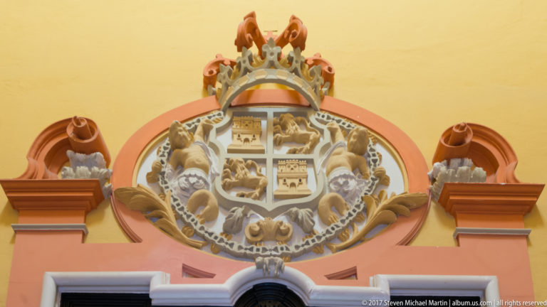 Polychrome crowned coat of arms with heraldic animals and castle motifs mounted above an arched opening on a yellow-and-rose wall.