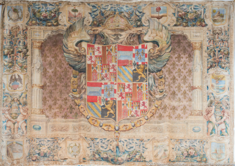 Large tapestry centered on a multi-field heraldic shield with castles, lions, stripes, surrounding scrollwork, figures, and decorative borders.