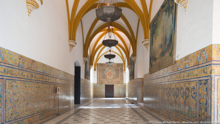 Long vaulted corridor with blue-and-yellow patterned tile dadoes, ochre ceiling ribs, hanging lanterns, and a tapestry on the right wall.