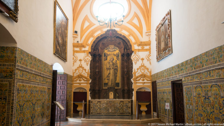 Narrow chapel with blue-and-yellow tiled walls, ochre-and-white vaulting, chandelier, framed paintings, and a dark carved altarpiece at the far end.
