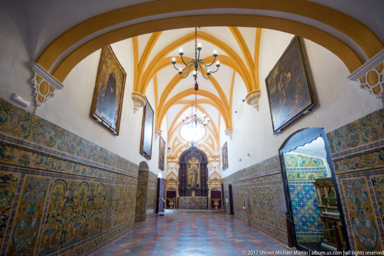 Axial view down a chapel with patterned tile walls, framed paintings, chandeliers, ochre vault ribs, and a dark altarpiece at the end.