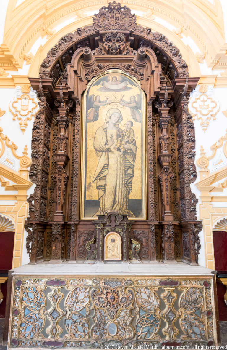 Carved dark wooden altarpiece with a central religious image above an altar faced in intricate blue, white, and ochre patterned tiles.