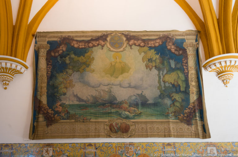 Large faded landscape tapestry mounted on a white wall between ochre ribs, above a narrow band of blue-and-yellow patterned tiles.