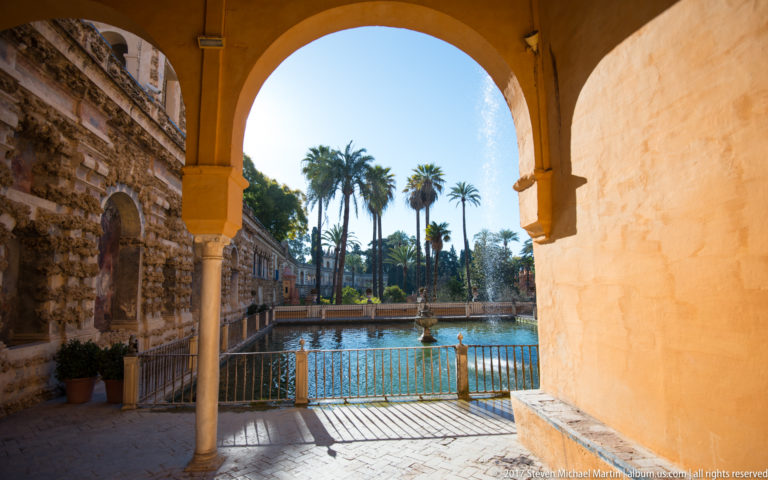 View through an ochre arch and column toward a blue garden pool, balustrade, fountain, palm trees, and dense greenery.